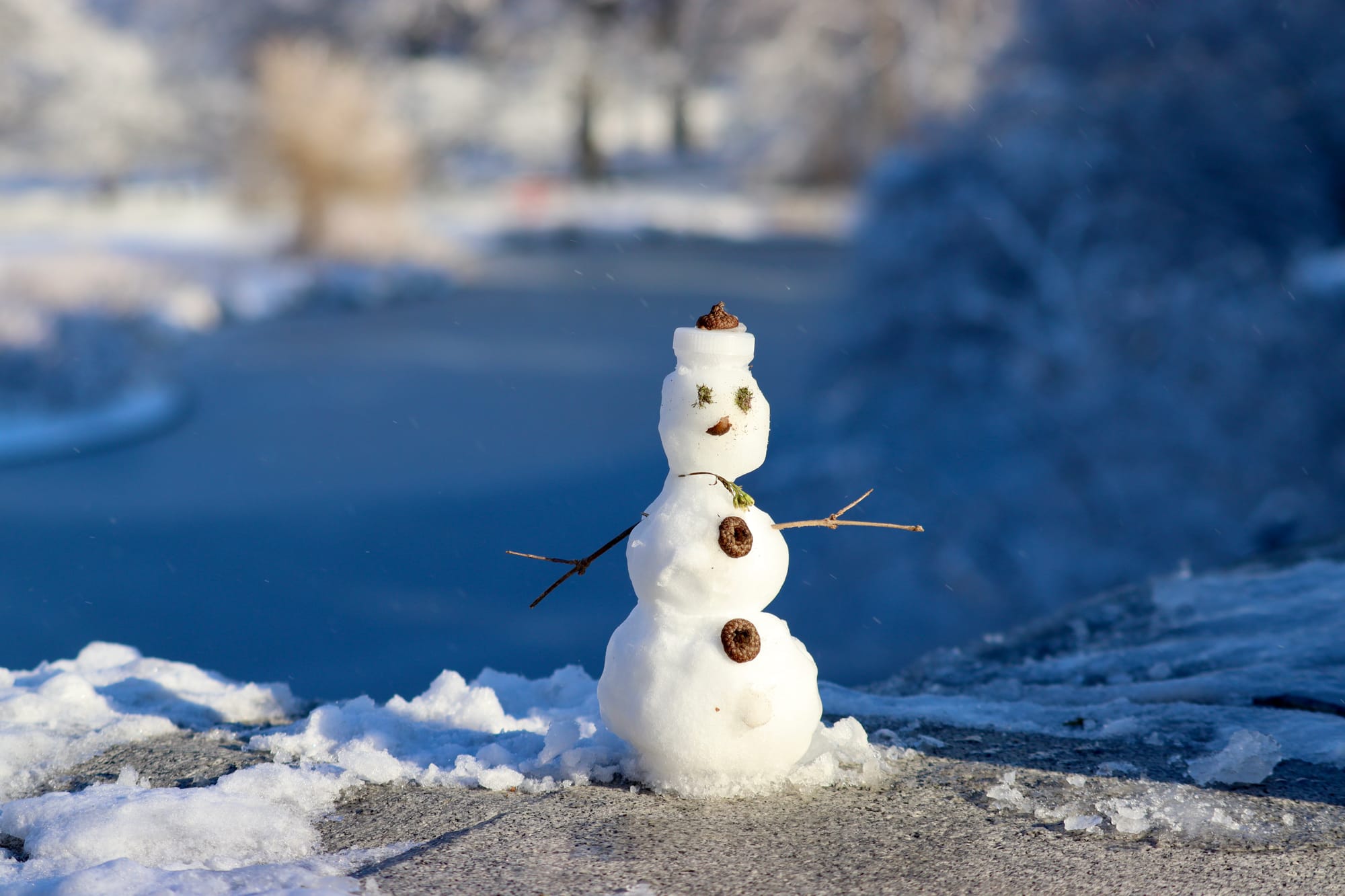 A tiny snowman sitting on a ledge with twig arms, acorn buttons and hat, and bits of greenery as a face and scarf