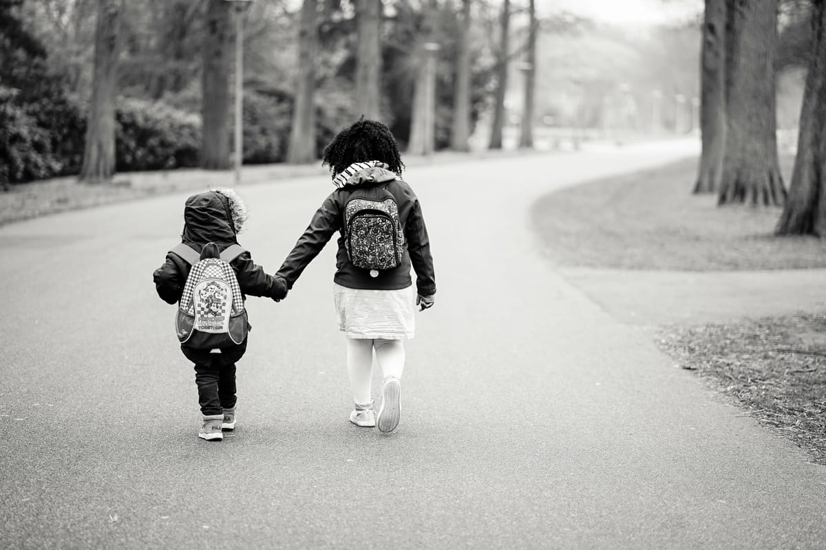 Photo of an older child holding a younger child's hand, viewed from behind, walking down a road