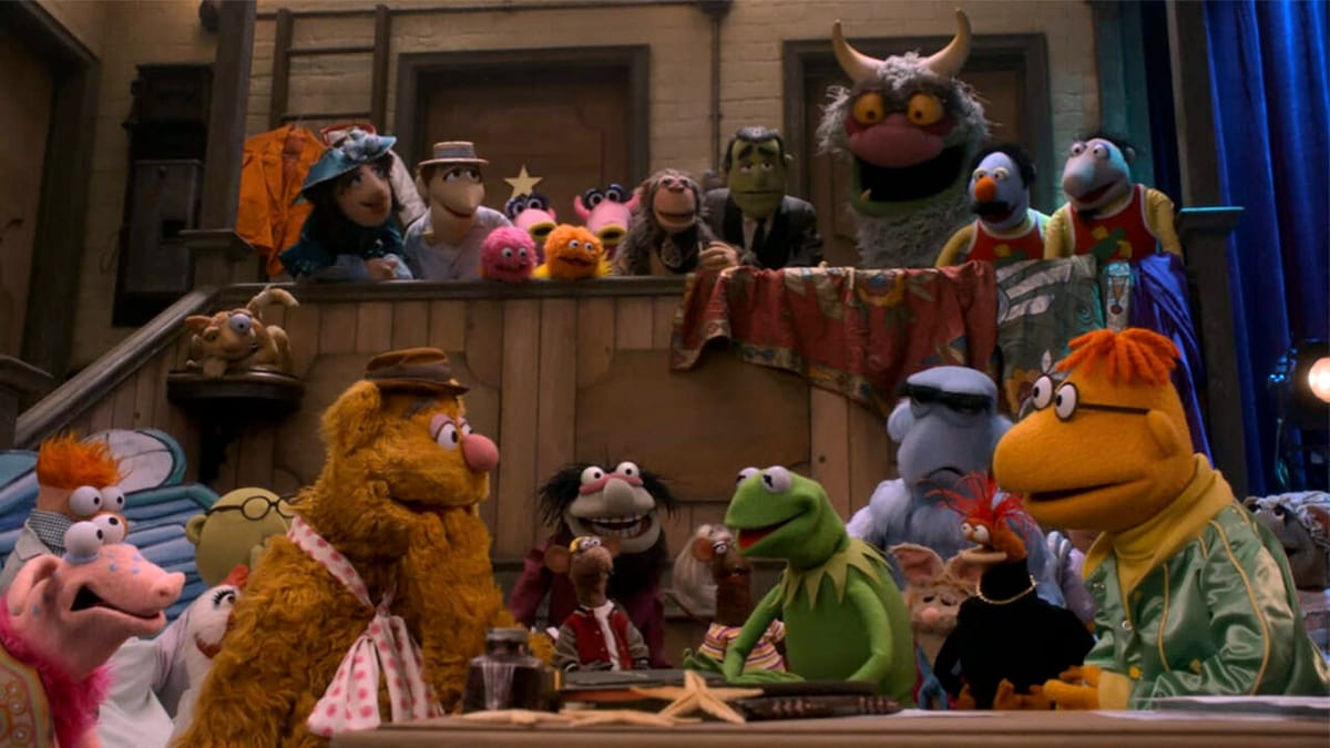 Muppet characters stand in a group back stage with Kermit and Fozzie in the center.