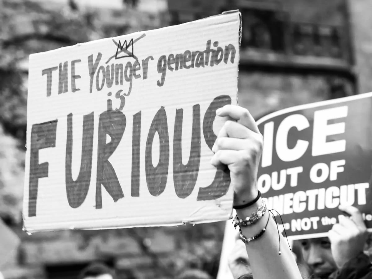 Black and white photo of a sign reading, "The Younger generation is FURIOUS," held overhead. The person holding the sign is wearing multiple beaded bracelets.