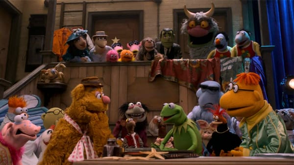 Muppet characters stand in a group back stage with Kermit and Fozzie in the center.